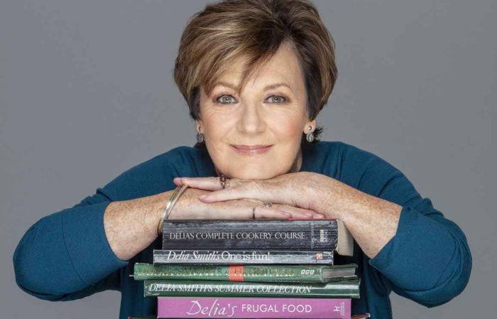 Delia Smith leans her chin on a stack of cookbooks, including titles like Delias Complete Cookery Course and Delia Smiths Christmas. She gazes directly at the camera with a neutral expression. The background is grey.