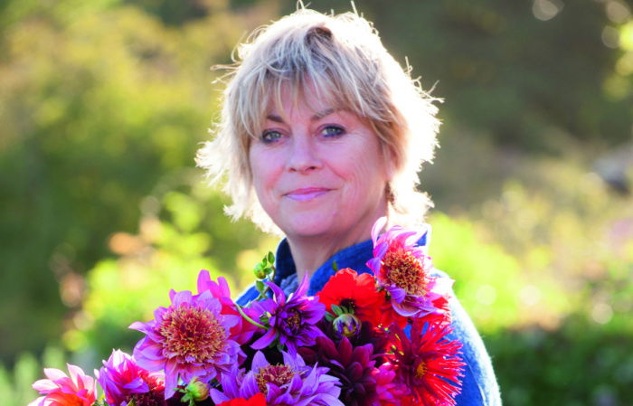 Sarah Raven stands outdoors, smiling and holding a colorful bouquet of flowers. The background is a green, sunlit garden.