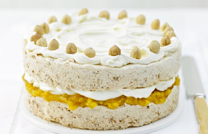 A layered cake with icing topped with whole hazelnuts. The middle layer contains a yellow filling, possibly fruit-based, creating a contrast with the beige cake. The cake is presented on a white surface with a knife beside it.