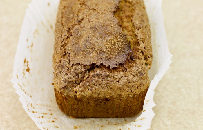 A loaf of banana bread with a slightly cracked top rests in a white paper liner on a light brown surface. The top has a textured, sugary crust.