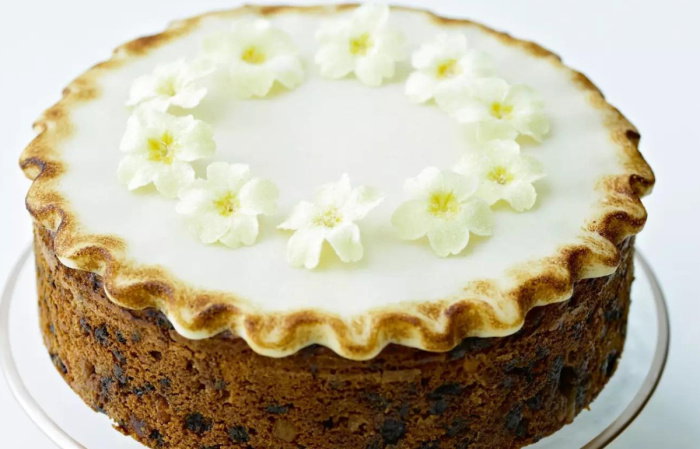 An Easter Simnel Cake on a glass cake stand decorated on the top with icing and little yellow and white flowers, next to a white plate of more white and yellow flowers and a gold ribbon next to it
