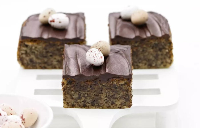Three chocolate-topped cake squares on a white serving spatula, each decorated with small speckled mini eggs. A bowl of additional eggs is in the foreground on a white surface.