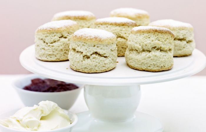 A white china cake stand with scones on top, two white dishes one of cream and one of jam and two teaspoons at the side