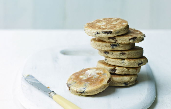 A stack of toasted cakes with raisins sits on a white marble board. A small butter knife with a light yellow handle rests beside the stack. The background is a soft white, creating a minimalist and airy setting.