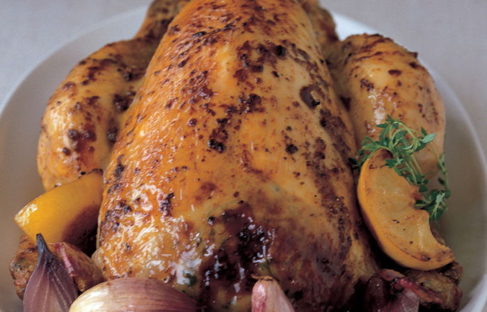 A picture of a roasted chicken in a dish surrounded by lemon wedges, thyme and garlic