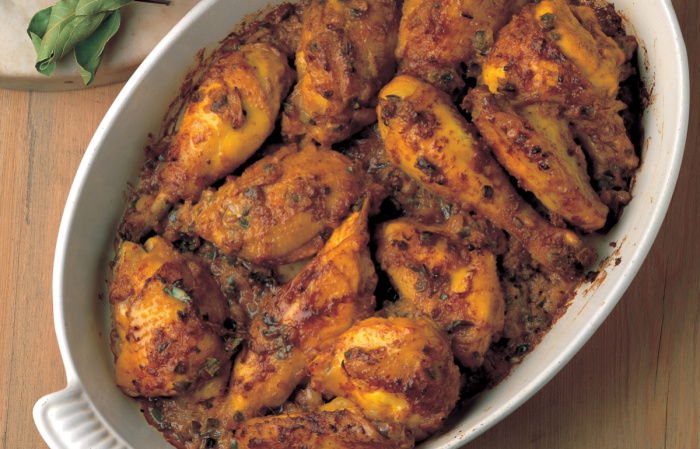 A white oval dish with pieces of spiced cooked chicken, a pestle and mortar next to it with ground spices and a plate of rice