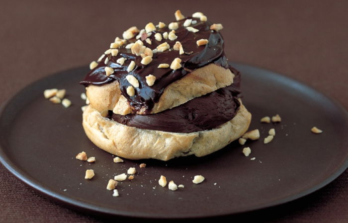 A choux bun split in half horizontally and filled and topped with a chocolate mousse mixture and coated with chocolate icing and chopped nuts.