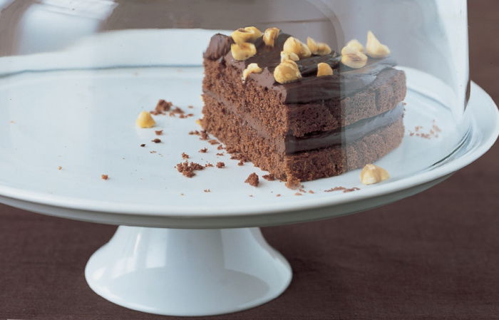 A slice of chocolate cake on a white cake stand with a glass lid