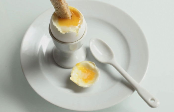a white china egg cup on a white plate, with a soft boiled egg opened and a solder being dipped in the yolk