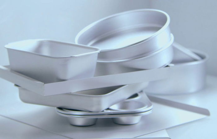 A stack of various silver baking pans, including round, rectangular, and muffin shapes, arranged randomly against a light grey background.