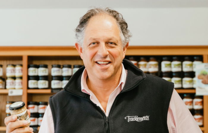 Guy Tullberg smiling while holding a jar, wearing a black vest with a logo. Shelves with numerous jarred products are visible in the background.