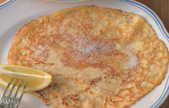 A picture of a cooked pancake covered in sugar on a white plate with a wedge of lemon on a fork resting on the side