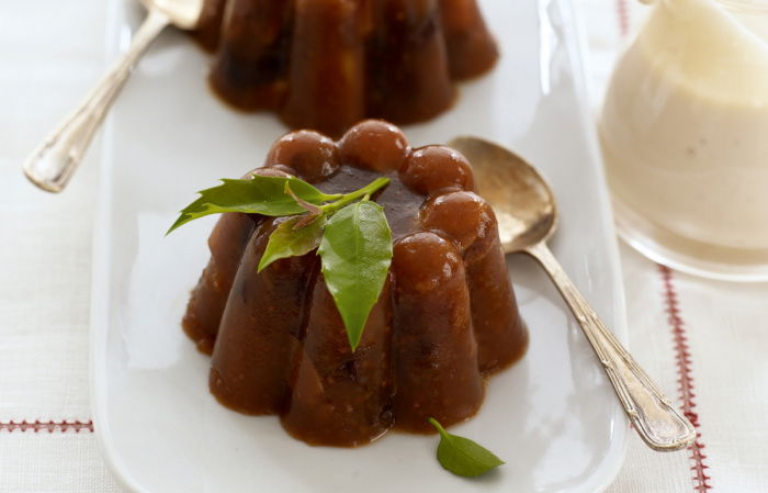 two pretty chrismas puddings on a whilte place with a spoon resting on the side and a jug of cream