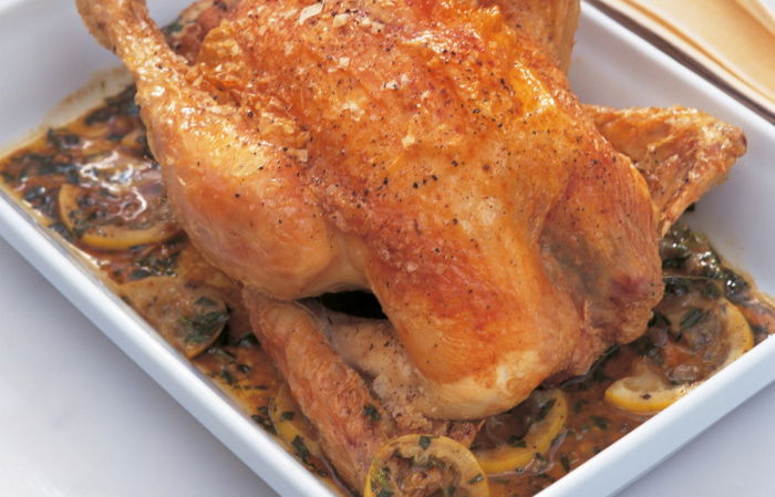 A cooked roast chicken in a white oven dish surrounded by the juices and slices of lemon