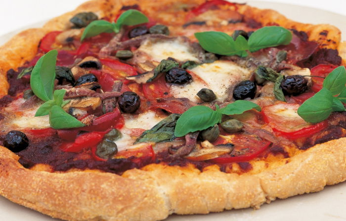 A cooked pizza on a white plate with a topping of basil, tomato, cheese and olives