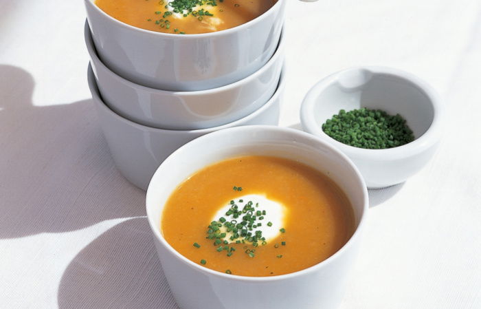 Four white soup bowls with two full of root vegetable soup, a white spoon resting on the top and a bowl of chopped parsley