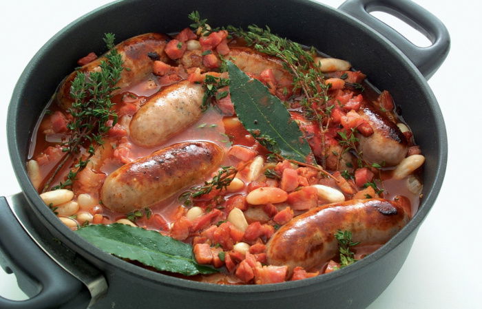 A picture of cooked cassoulet in a black casserole showing sausages, beans, bay leaves and tomatoes
