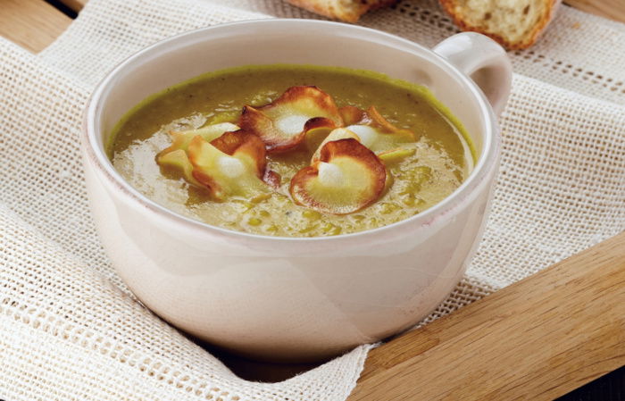 A bowl of parsnip soup in a white mug, with parsnip crisps on the top, sittin on a wooden tray with a roll of white bread next to it