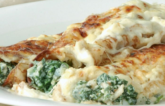 A picture of cooked pancake cannelloni on a white plate with spinach and a cheese sauce