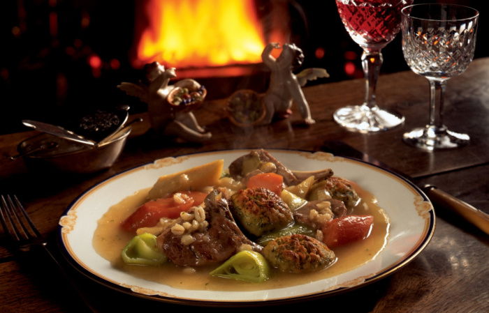 a picture of a table set with a plate of irish stew and a glass of wine witih a fire glowing in the background