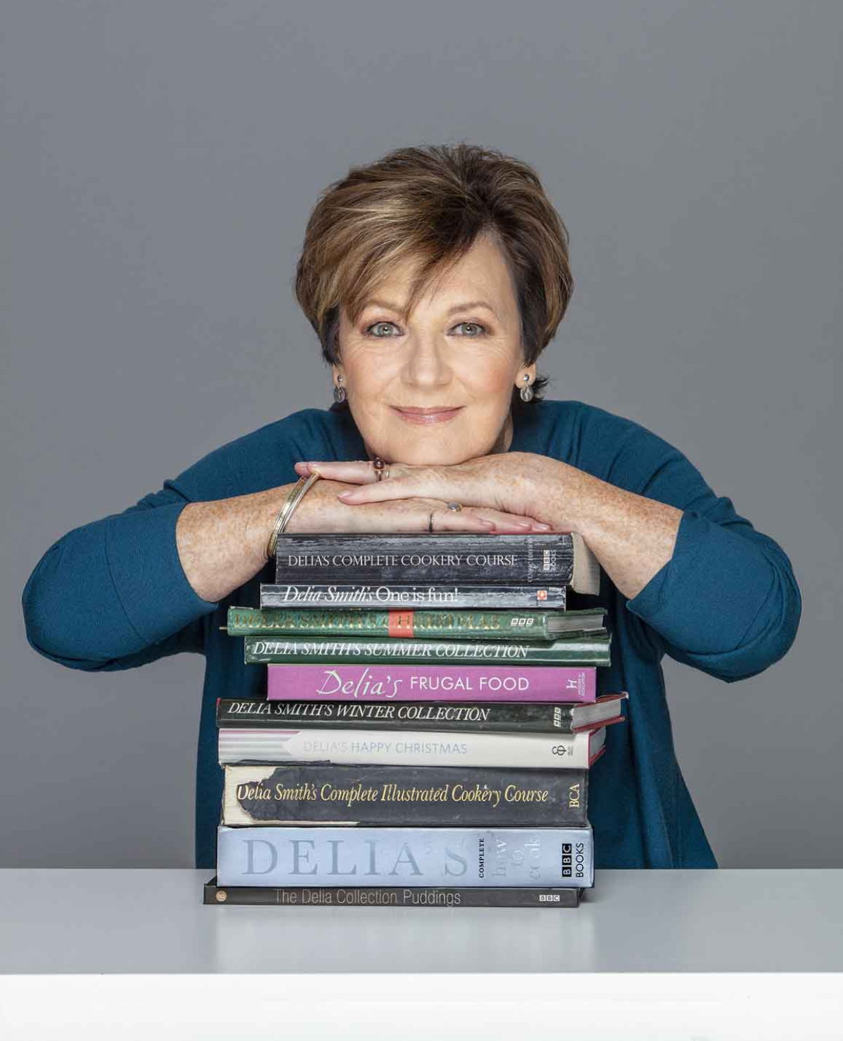 Delia Smith leans her chin on a stack of cookbooks, including titles like Delias Complete Cookery Course and Delia Smiths Christmas. She gazes directly at the camera with a neutral expression. The background is grey.