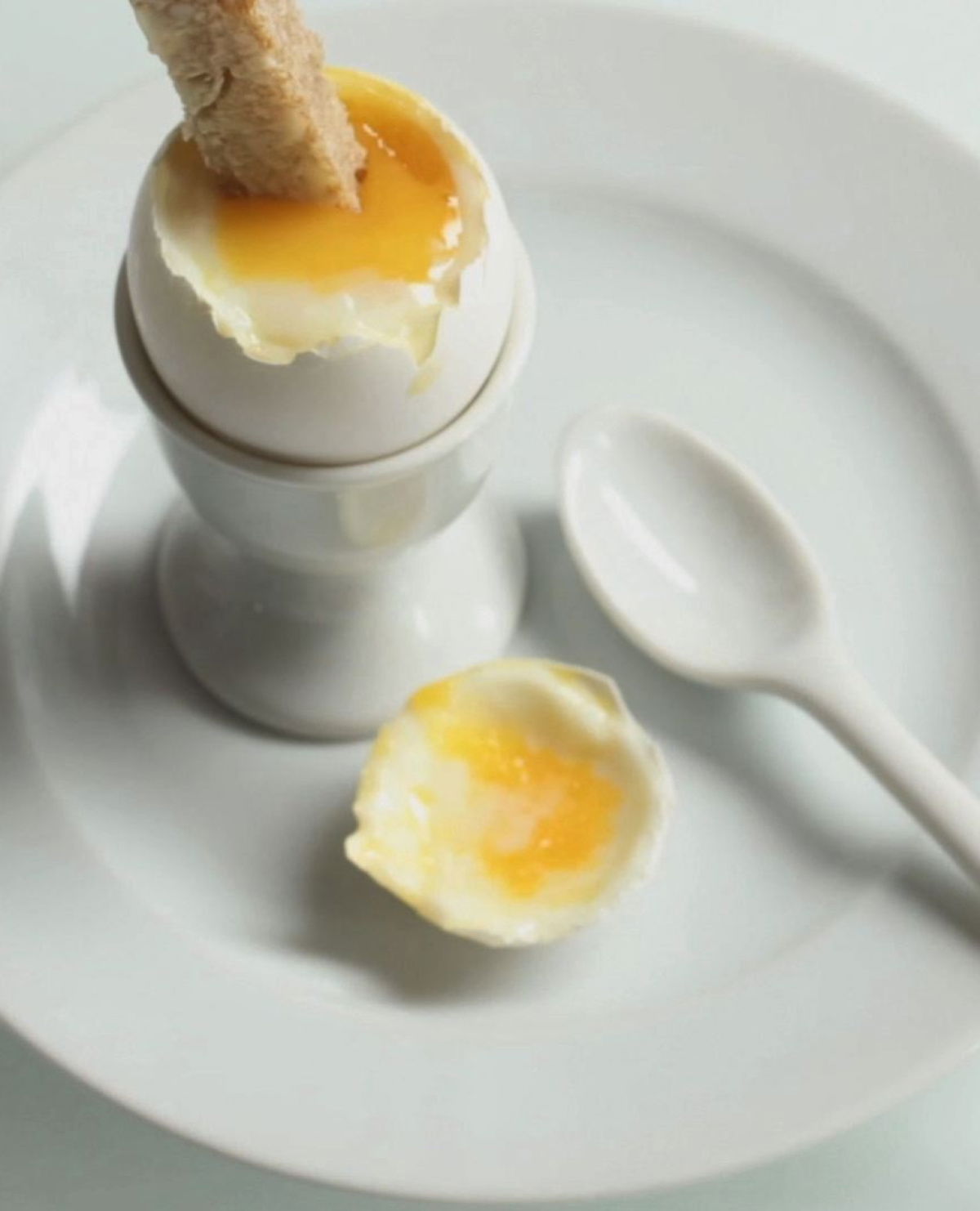 a white china egg cup on a white plate, with a soft boiled egg opened and a solder being dipped in the yolk
