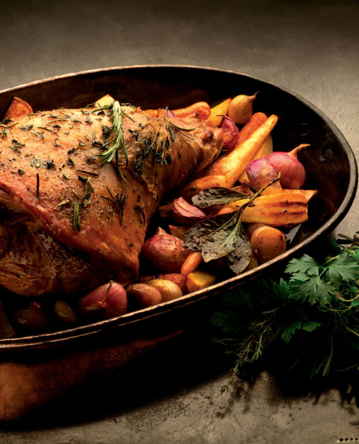 a cooked leg of lamb in a casserole dish with carrots and rosemary