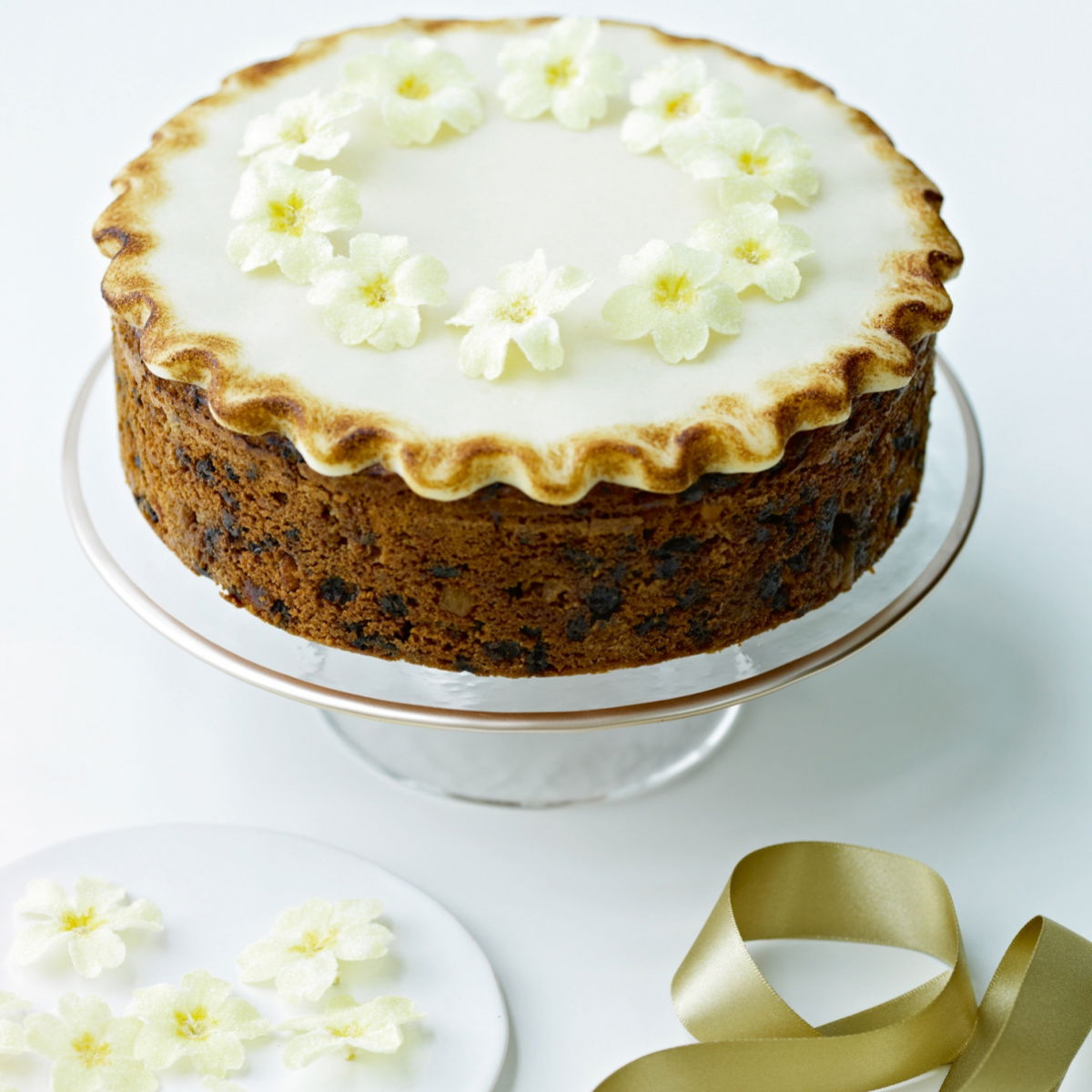 An Easter Simnel Cake on a glass cake stand decorated on the top with icing and little yellow and white flowers, next to a white plate of more white and yellow flowers and a gold ribbon next to it