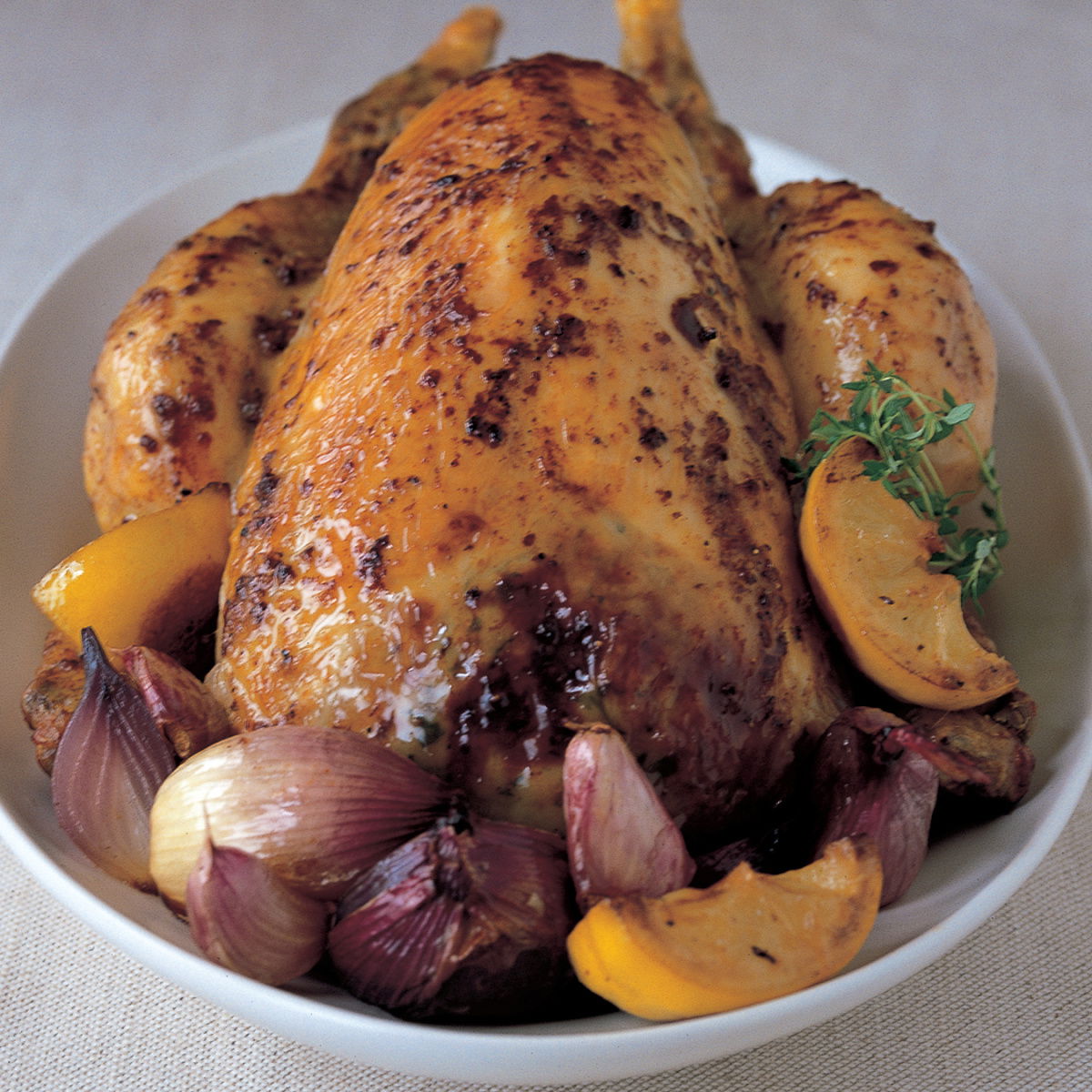 A picture of a roasted chicken in a dish surrounded by lemon wedges, thyme and garlic
