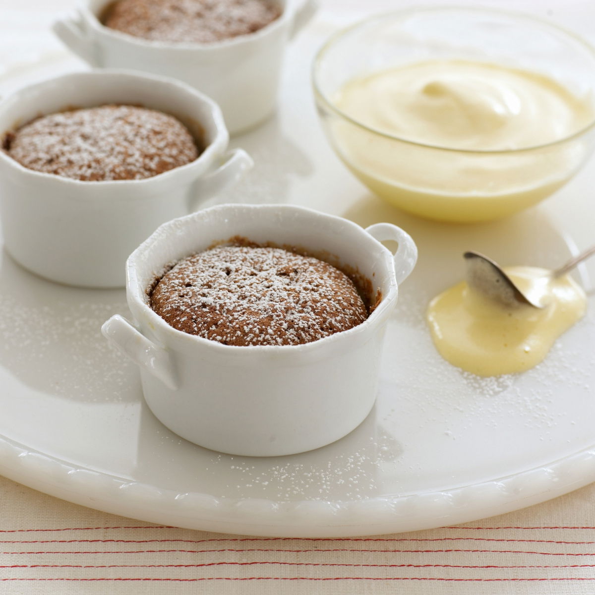 Three little white dishes on a white plate, with souffles dusted with icing sugar and a glass bowl of sabayon on the side