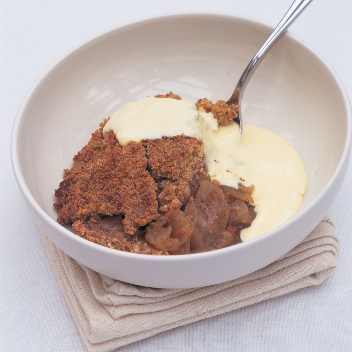 A white bowl of apple crumble with custard poured over the top and a spoon resting on the side