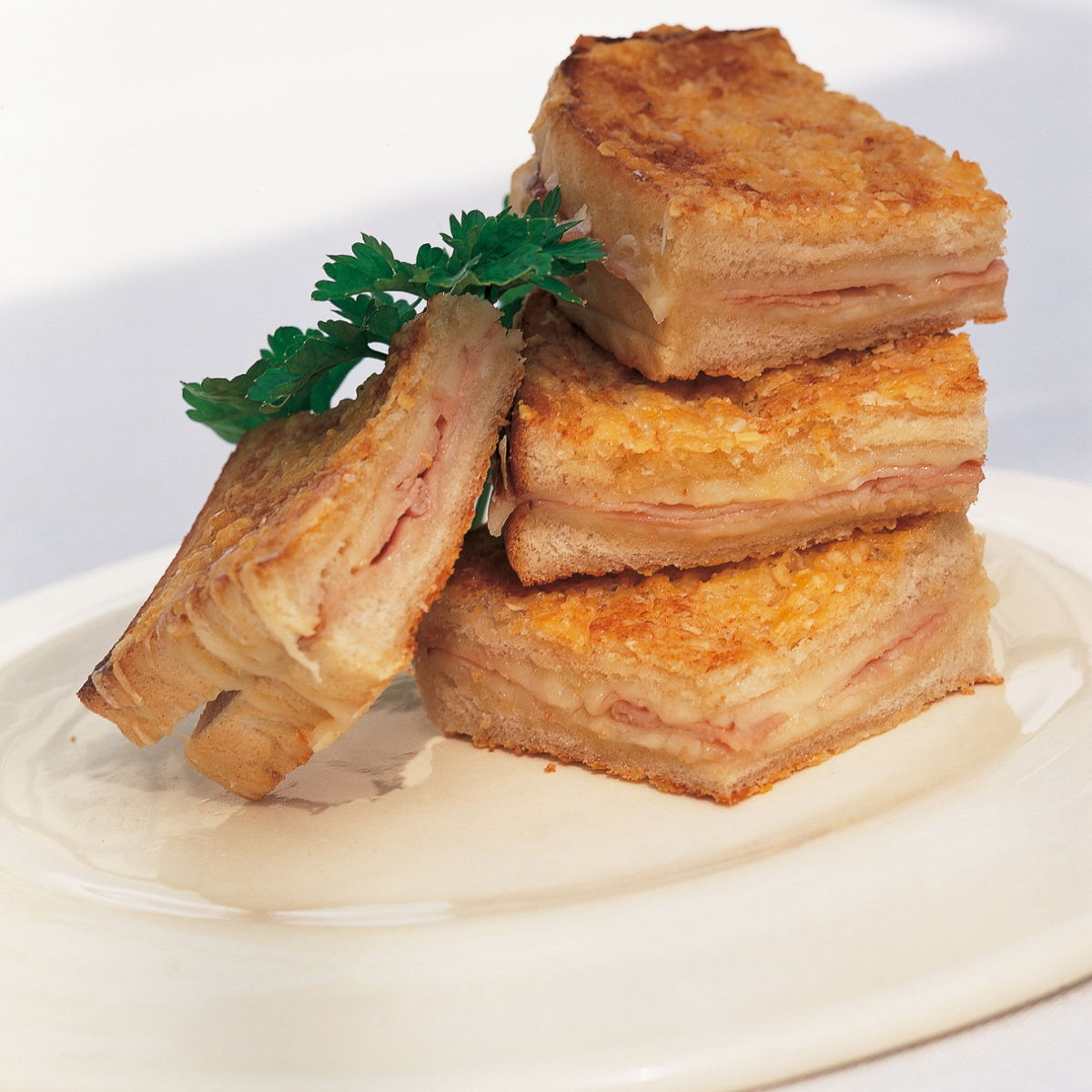Stacked croque monsieur sandwiches on a white plate, garnished with parsley. The sandwiches have layers of melted cheese and ham, with a golden, crispy crust.