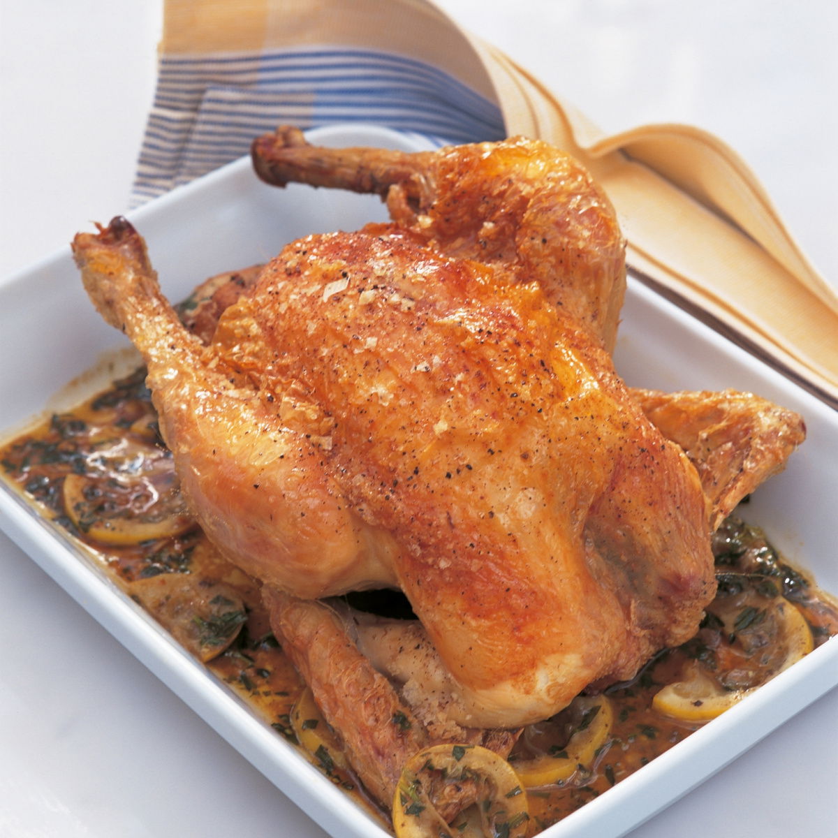 A cooked roast chicken in a white oven dish surrounded by the juices and slices of lemon