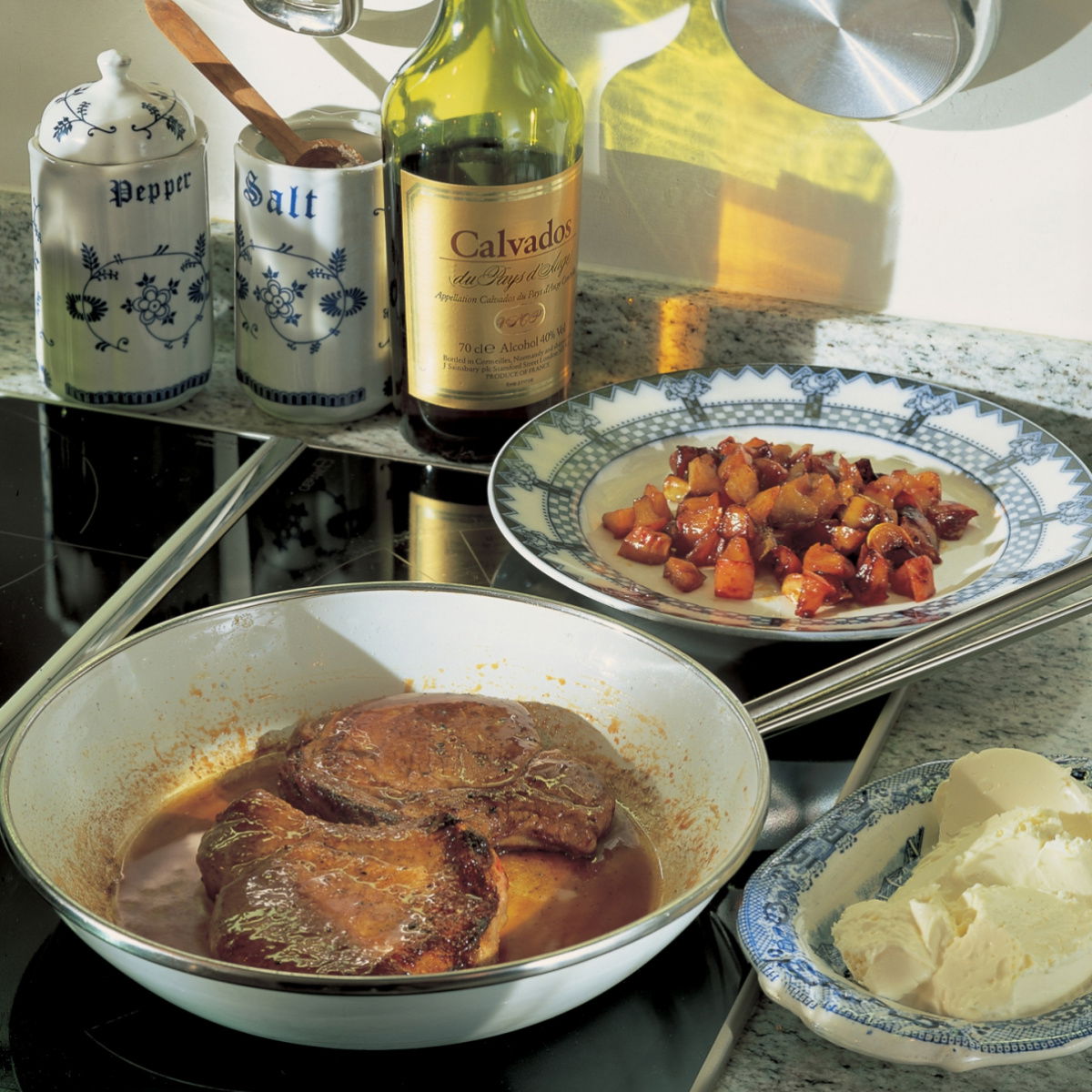 A hob with a white frying pan containing the cooking pork chops, with a plate of cooked chopped apples and shallots and a bowl of creme fraiche next to it
