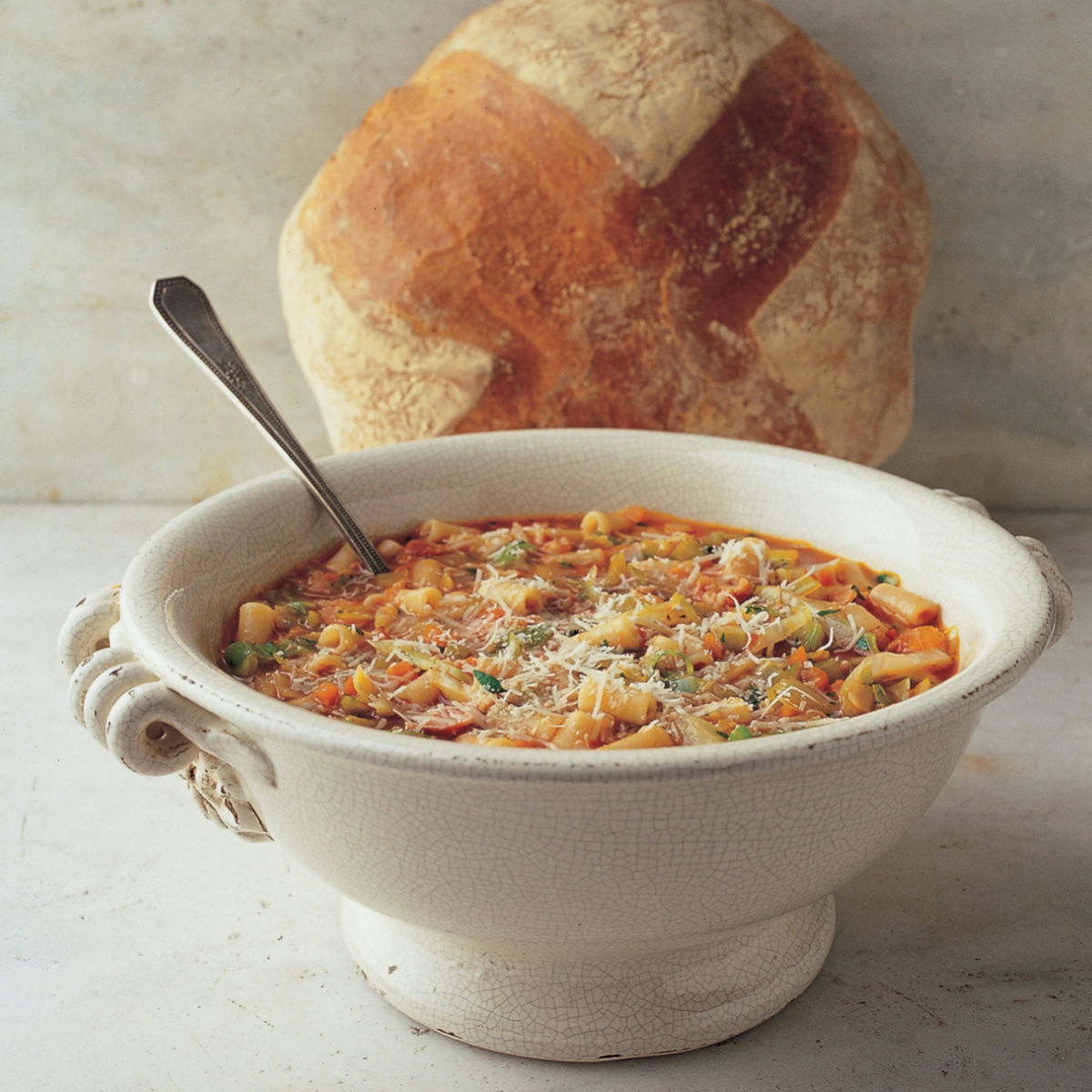 A white bowl with pasta soup garnished with herbs and cheese, with a spoon inside. A round loaf of bread is placed behind the bowl on a marble surface.