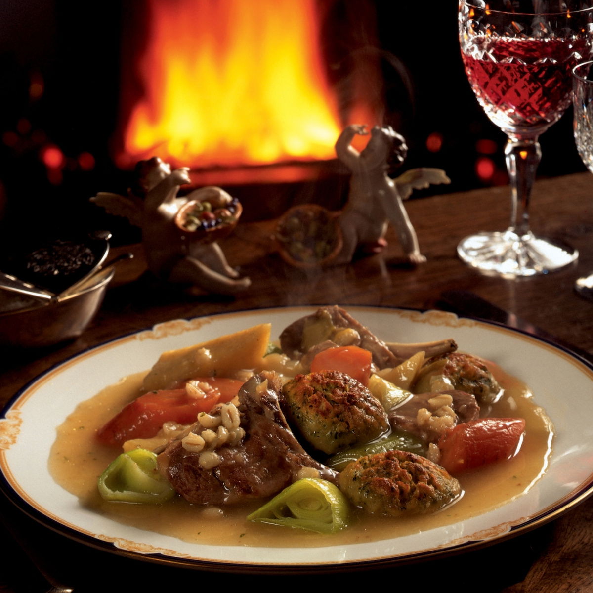 a picture of a table set with a plate of irish stew and a glass of wine witih a fire glowing in the background