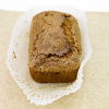 A loaf of banana bread with a slightly cracked top rests in a white paper liner on a light brown surface. The top has a textured, sugary crust.