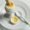 a white china egg cup on a white plate, with a soft boiled egg opened and a solder being dipped in the yolk