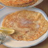 A picture of a cooked pancake covered in sugar on a white plate with a wedge of lemon on a fork resting on the side