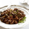 A white plate of cooked slices of venison, mushrooms and bacon in a sauce with some garnish on the side and a spoon resting on the rim