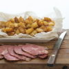 A picture of diced sauteed potatoes in a white dish sitting on a wooden board with sliced ham and a knife on the side