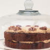 A layered cake with frosting and walnuts on top, displayed under a glass dome.