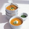 A picture of four white soup bowls with two full of root vegetable soup, a white spoon resting on the top and a bowl of chopped parsley