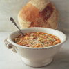 A white bowl with pasta soup garnished with herbs and cheese, with a spoon inside. A round loaf of bread is placed behind the bowl on a marble surface.