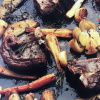A rustic roasted dish featuring pieces of meat, whole garlic cloves, carrot slices, and sprigs of herbs on a dark baking tray. The ingredients are browned and glistening, suggesting they were cooked in an oven.