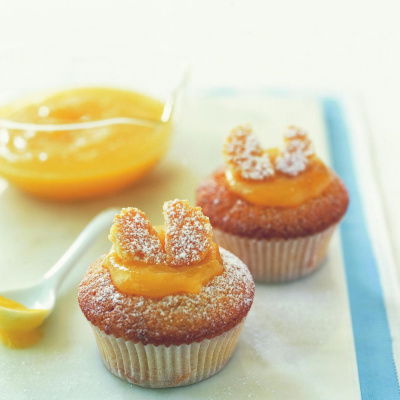 Two cupcakes topped with powdered sugar and slices resembling butterfly wings, placed on a white surface. A bowl of orange sauce and a spoon are in the background.