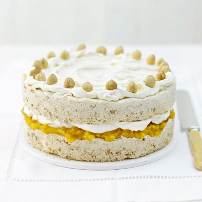 A layered cake with icing topped with whole hazelnuts. The middle layer contains a yellow filling, possibly fruit-based, creating a contrast with the beige cake. The cake is presented on a white surface with a knife beside it.