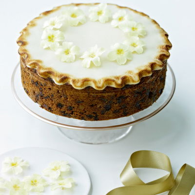 An Easter Simnel Cake on a glass cake stand decorated on the top with icing and little yellow and white flowers, next to a white plate of more white and yellow flowers and a gold ribbon next to it