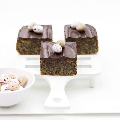 Three chocolate-topped cake squares on a white serving spatula, each decorated with small speckled mini eggs. A bowl of additional eggs is in the foreground on a white surface.