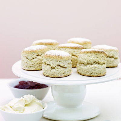 A white china cake stand with scones on top, two white dishes one of cream and one of jam and two teaspoons at the side